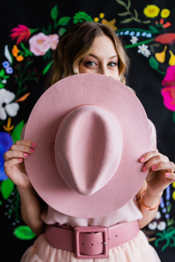 Model peeking over hat in front of painted floral backdrop on black paper
