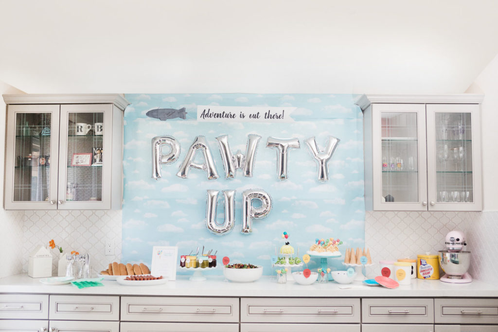 Food and dessert display with blue sky backdrop and balloons that spell "Pawty Up"