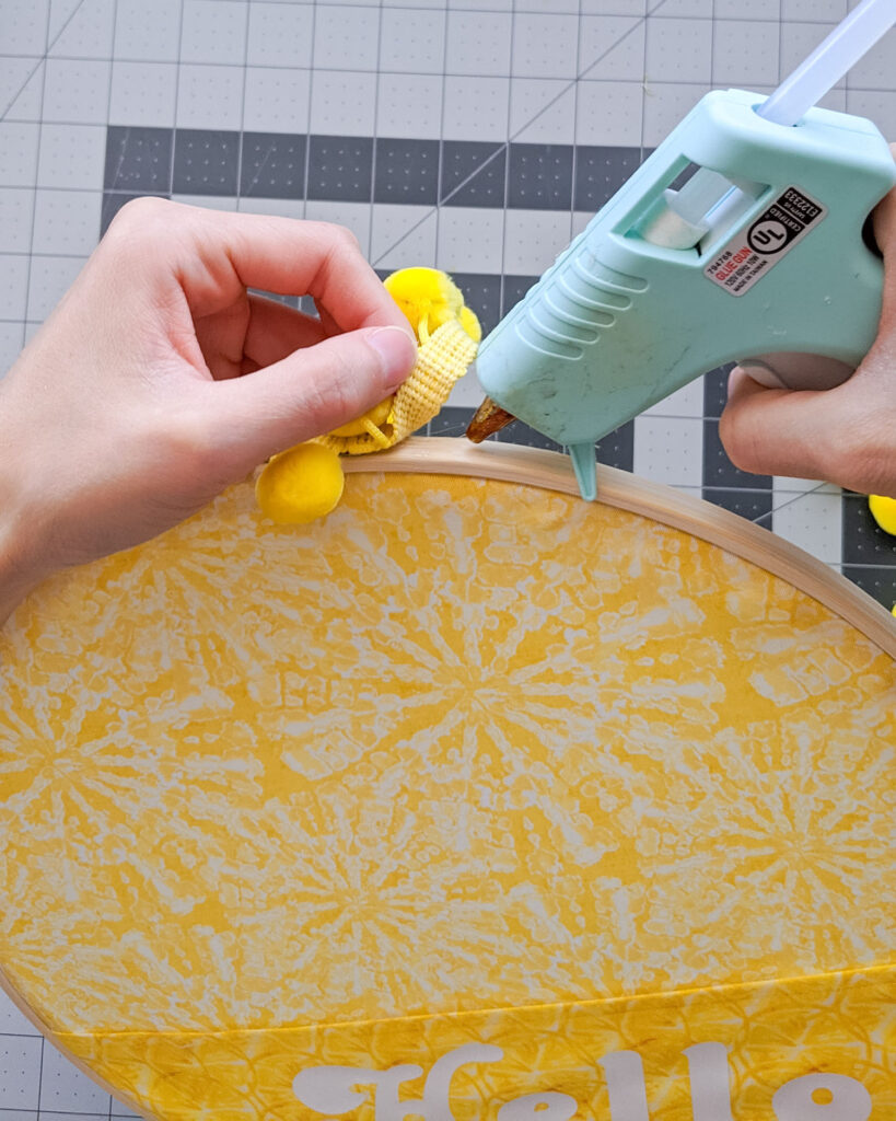 Hot gluing yellow pom pom trim around embroidery hoop decor