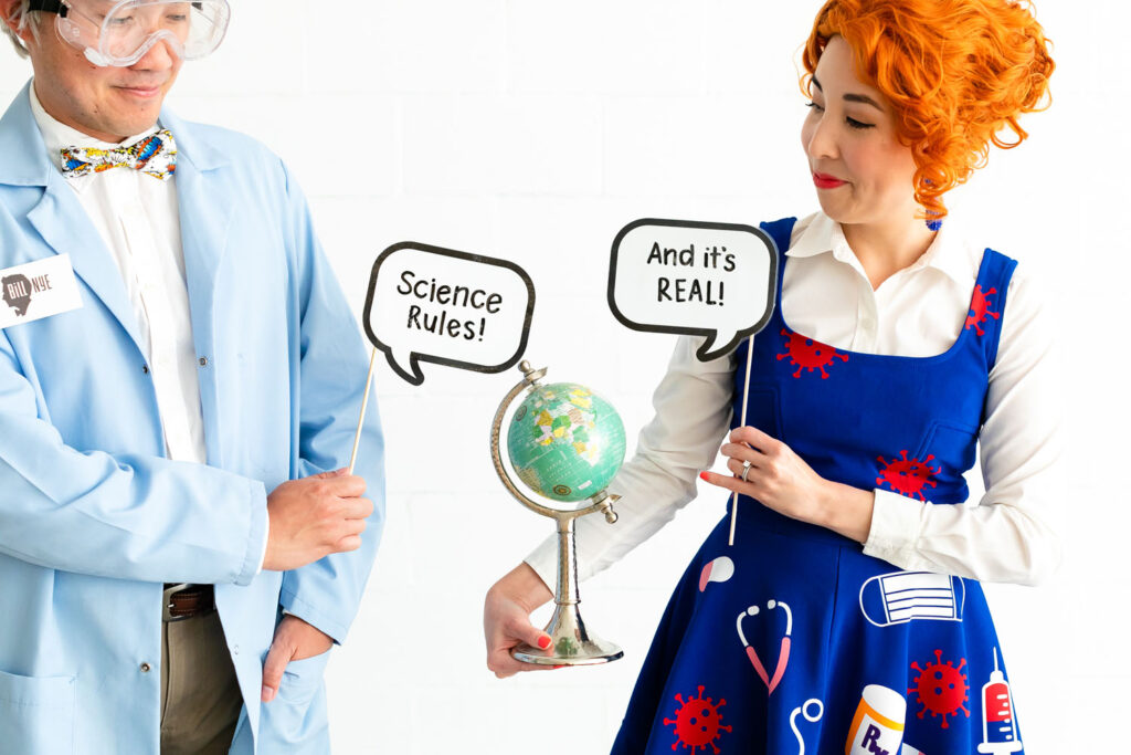 Couple dressed as Bill Nye and Ms. Frizzle holding signs that say "Science Rules" "And it's Real". 