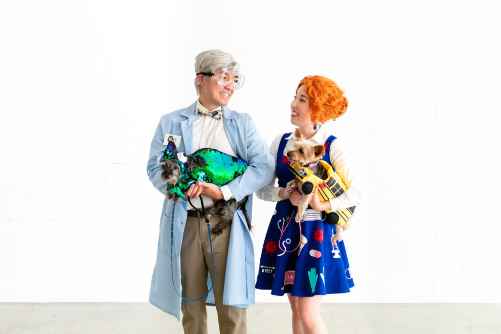 Couple dressed as Bill Nye and Ms. Frizzle, holding their dogs dressed as the Magic School Bus and Liz the chameleon.