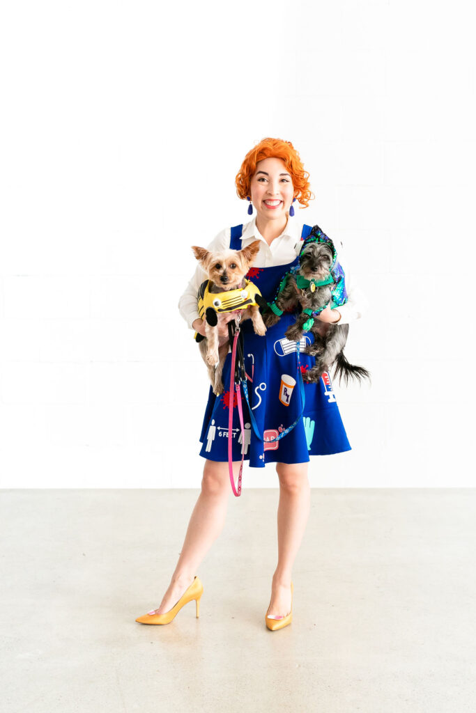 Craft blogger dressed in Ms. Frizzle costume. She is holding two dogs dressed as the Magic School Bus and Liz the chameleon.