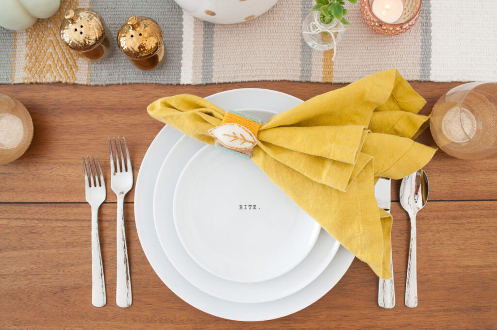 Close up of Thanksgiving table place setting featuring white plates, mustard napkin with yarn napkin ring, silver flatware and a gold glitter wine glass.
