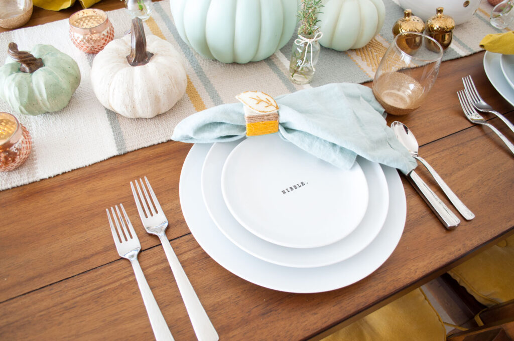 Thanksgiving table showcasing a single place setting. There are three white plates stacked. The small top plate says "nibble". A blue linen napkin is placed on top with a handmade napkin ring.