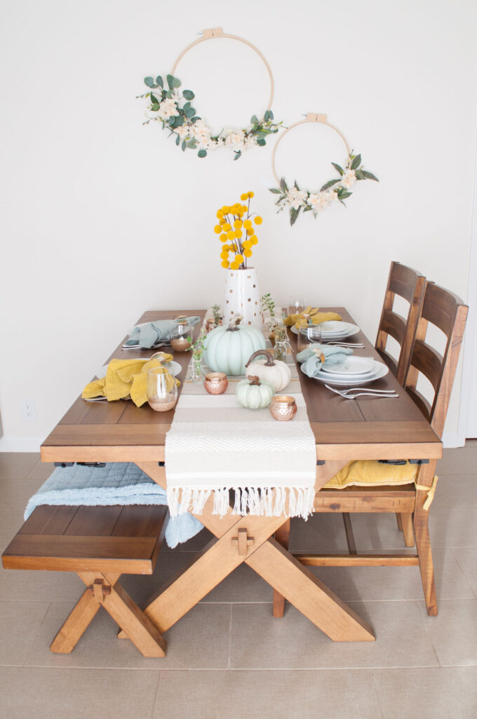 Wood table with cross-legged base, bench and two chairs. It's decorated for Thanksgiving with aqua and mustard decor.