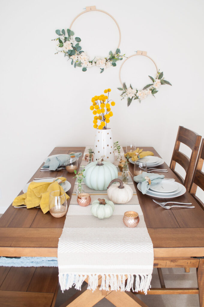 Side view of casual Thanksgiving table. A woven runner with tassels is draped down the middle and covered with fall decor.