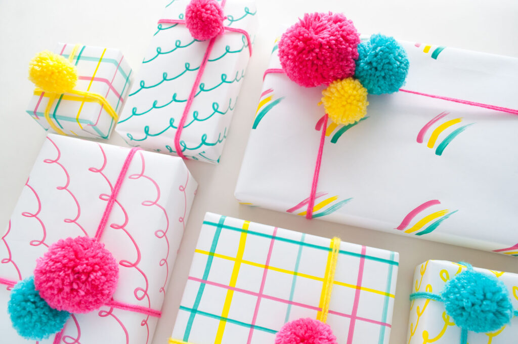 Close up of hand drawn gift wrap. The three designs are loops, grid pattern and rainbow brush strokes. The designs are drawn with pink, yellow and teal paint markers. The gifts are wrapped with coordinating pom poms.