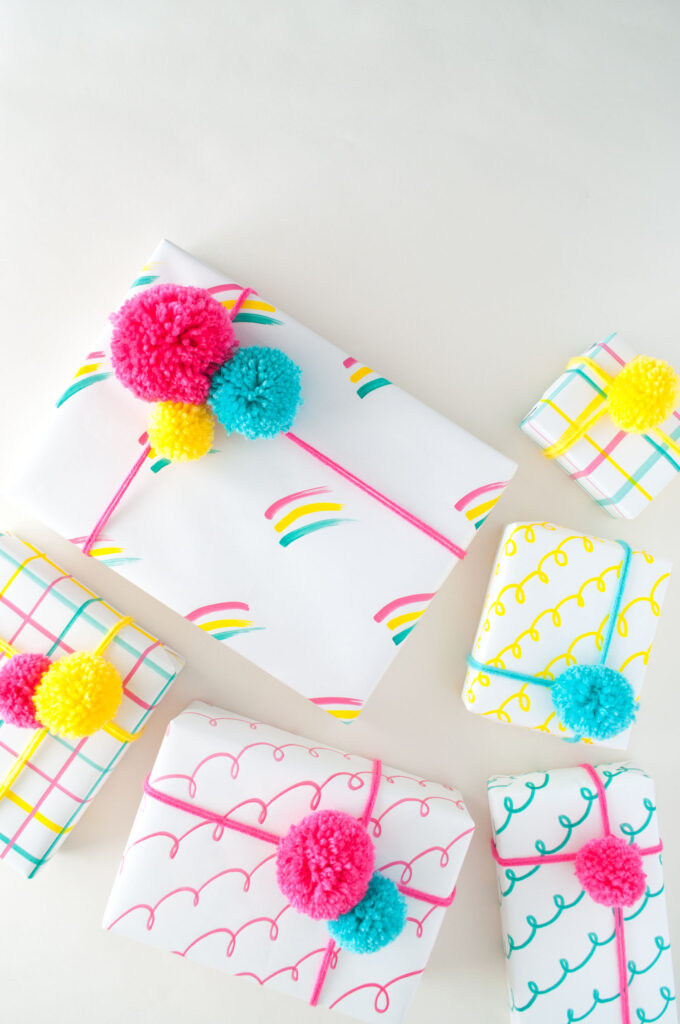 Hand drawn gift wrap using COLORSHOT markers. The designs are rainbows, loops and a grid pattern. The colors used are hot pink, yellow and teal. They're decorated with matching pom poms.
