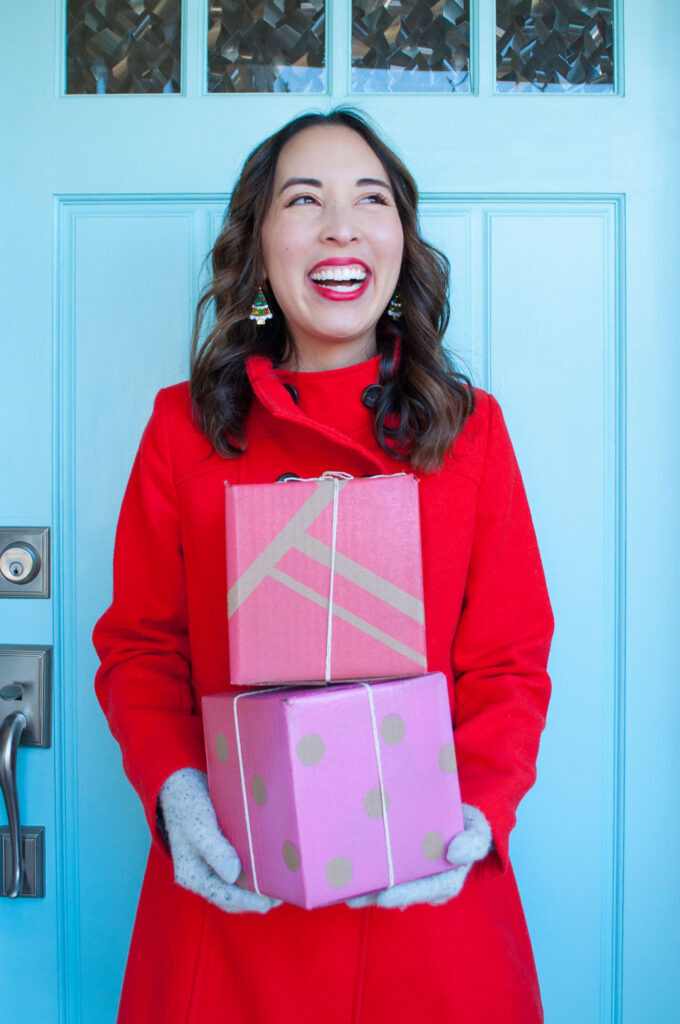 Blaire from Freshly Fuji holding the completed gift boxes. She's wearing a red coat and smiling, standing in front of a blue door. The gift boxes are blank cardboard boxes she upgraded with a paint resist technique.
