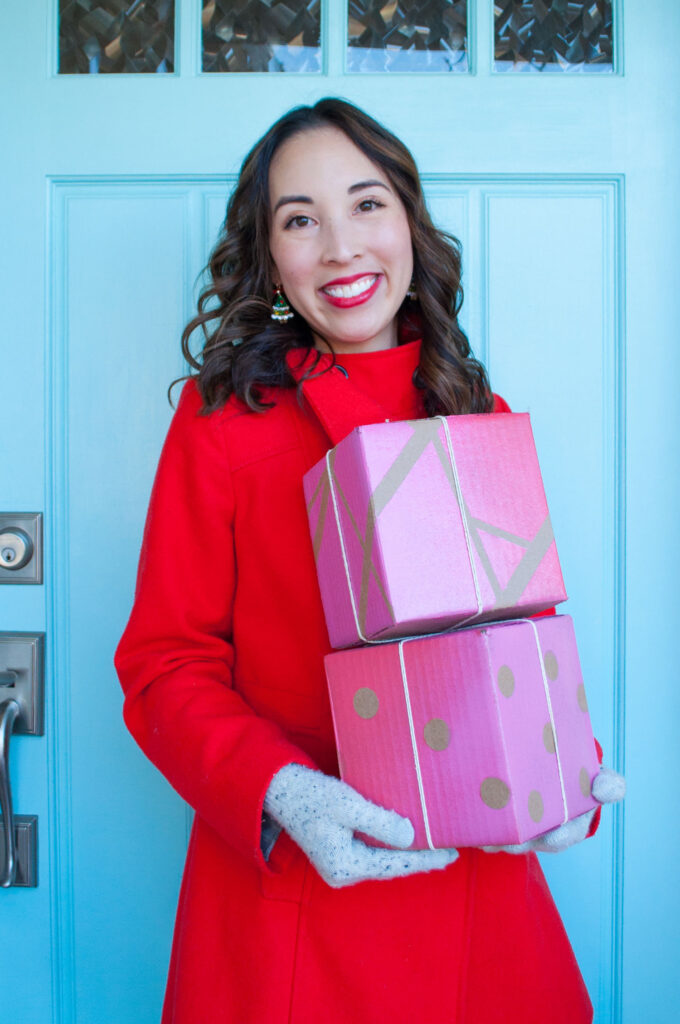 Blaire from Freshly Fuji holding the completed gift boxes. She's wearing a red coat and smiling, standing in front of a blue door. The gift boxes are blank cardboard boxes she upgraded with a paint resist technique.