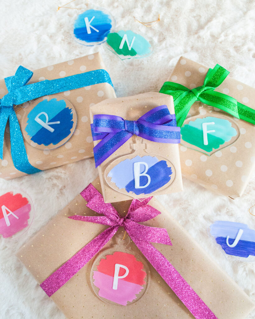 Gifts wrapped in brown paper showcasing the final holiday gift wrap hack: Painted acrylic gift tags with initials on them. The tags are painted in a color block fashion - half in one color, half in another color.