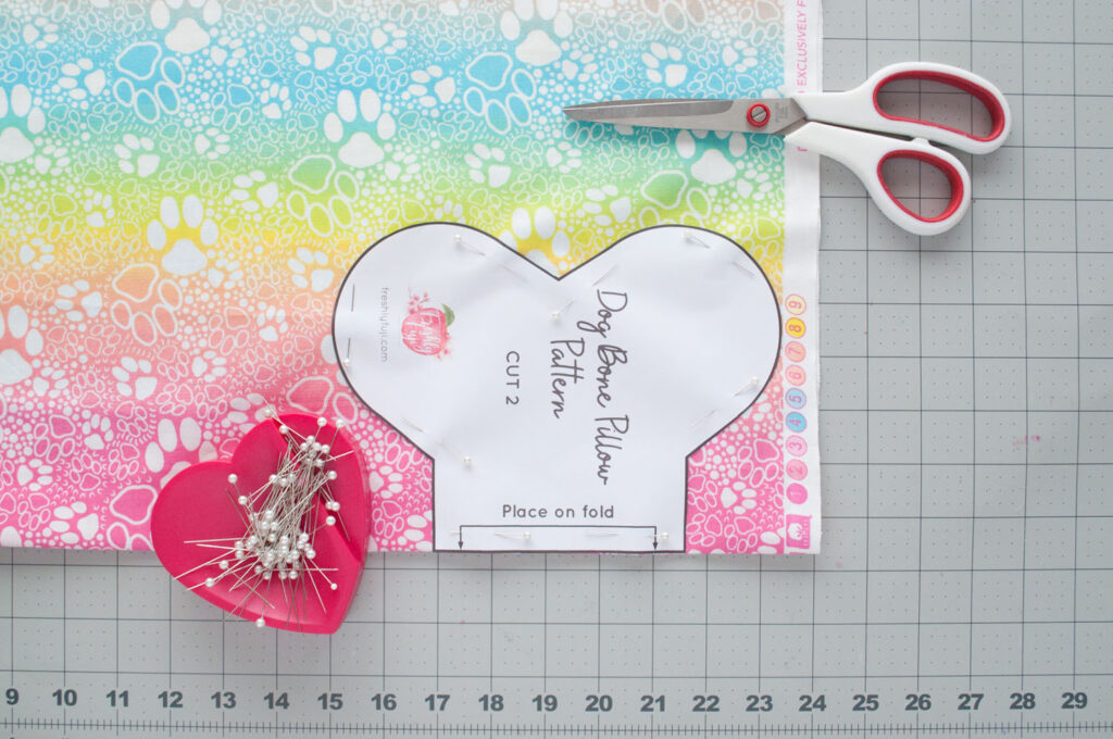 Dog bone pillow pattern is pinned onto the rainbow paw print fabric in preparation for cutting.