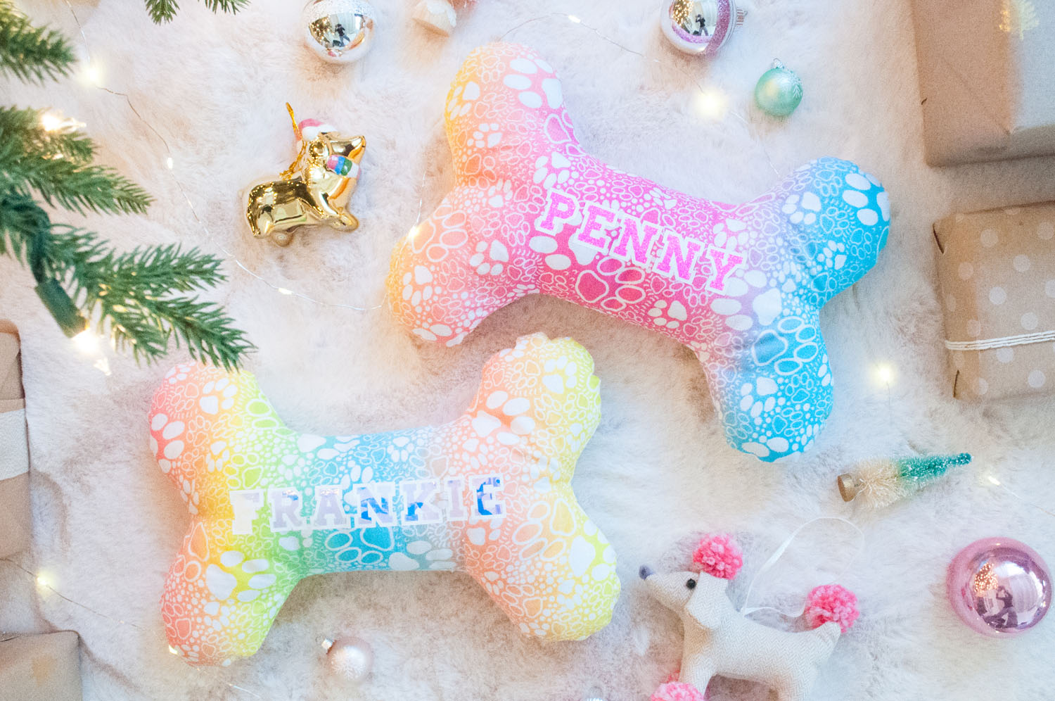 Photo of dog bone pillows. The fabric has a dog bone print on a gradient rainbow background. The pillows are personalized with the names Frankie and Penny in Iron-On material. They are placed on a fur background with ornaments and presents.