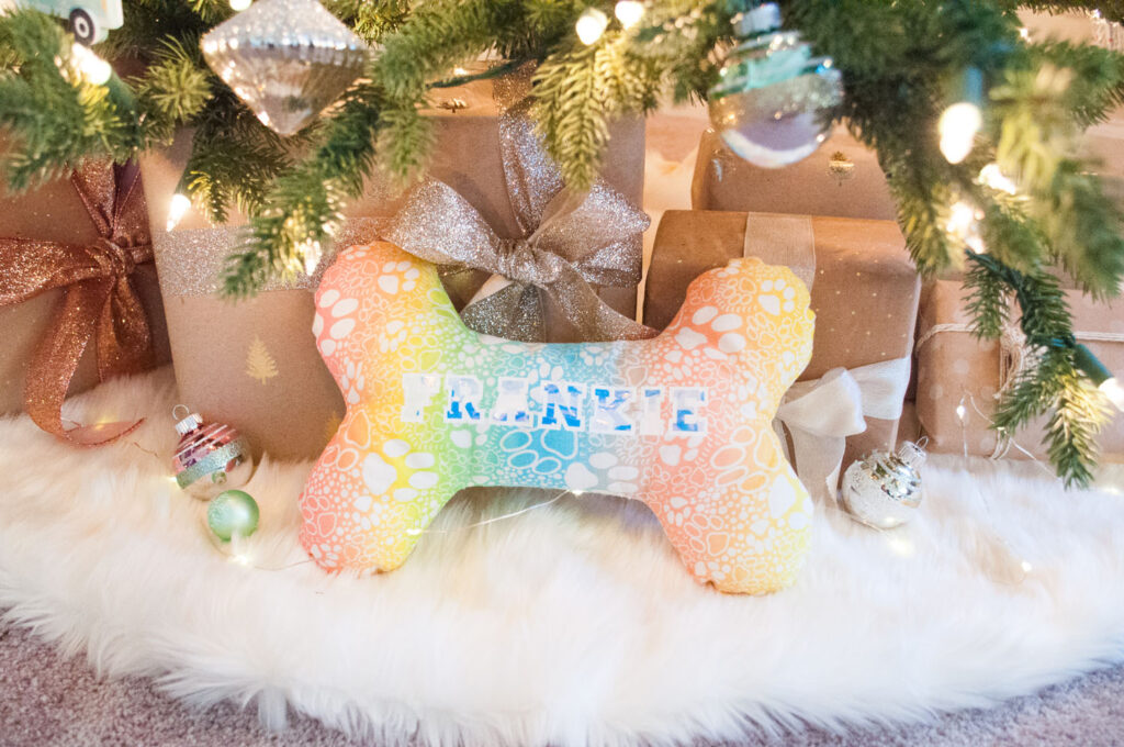 Close up of dog bone pillow. The fabric has a colorful gradient background and is covered in paw prints. The pillow is placed under a Christmas tree amongst presents and ornaments.