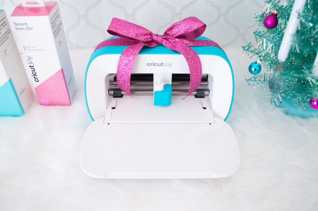 Close up of Cricut Joy machine with door open. There's a glitter fuchsia bow on top if it. To the left are two boxes of Smart iron-On and to the right is the base of an aqua tabletop tree.