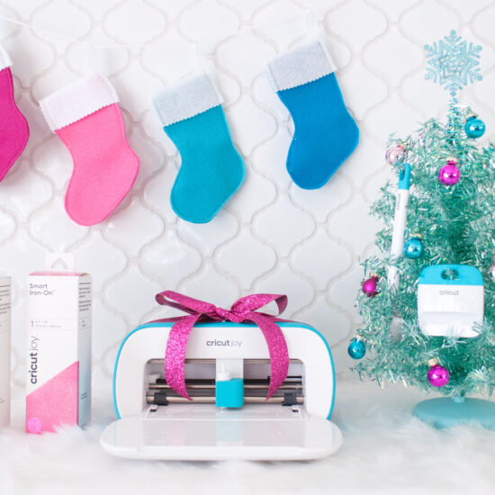 A Christmas scene with white tile backsplash. There are stockings hung on the top. At the bottom is two Smart Iron-On boxes, the Cricut Joy with a fuchsia bow and an aqua tabletop tree with Cricut tools hung from it.