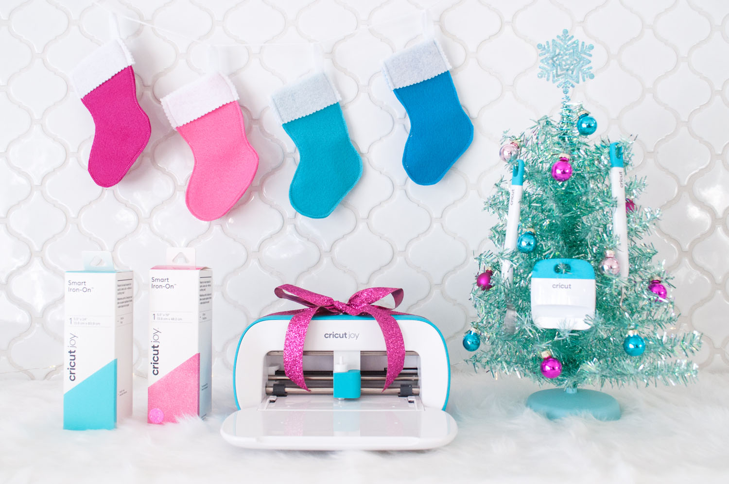 A Christmas scene with white tile backsplash. There are stockings hung on the top. At the bottom is two Smart Iron-On boxes, the Cricut Joy with a fuchsia bow and an aqua tabletop tree with Cricut tools hung from it.