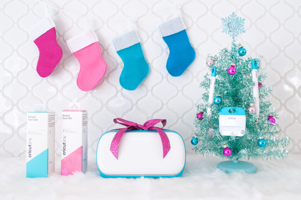 A Christmas scene with white tile backsplash. There are stockings hung on the top. At the bottom is two Smart Iron-On boxes, the Cricut Joy with a fuchsia bow and an aqua tabletop tree with Cricut tools hung from it.