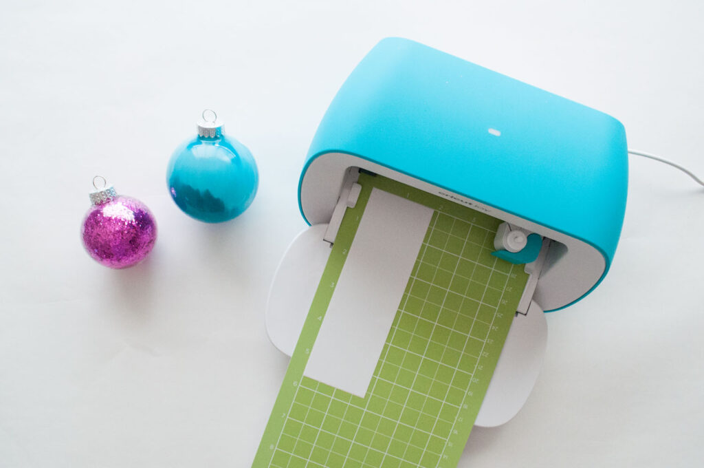 A Cricut Joy machine with the door open. There's a green StandardGrip mat loaded with a small piece of vinyl. There is a fuchsia glitter ornament and teal painted ornament on the left.