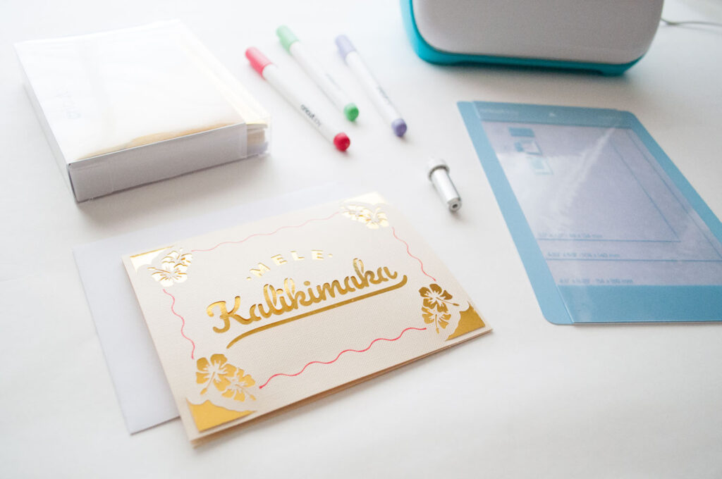 Close up of card made with Cricut Joy. It says "Mele Kalikimaka" in gold foil. There are also gold foil hibiscus on the cream colored card.