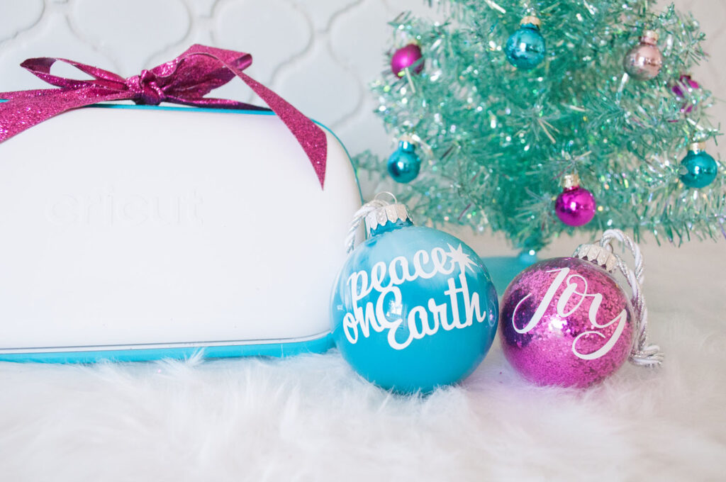 Two ornaments customized with vinyl phrases. The aqua ornament says "Peace on Earth" and the fuchsia glitter ornament says "Joy". To the left is the Cricut Joy with a fuchsia glitter bow. Behind the ornaments is an aqua Christmas tree decorated with mini ornaments.