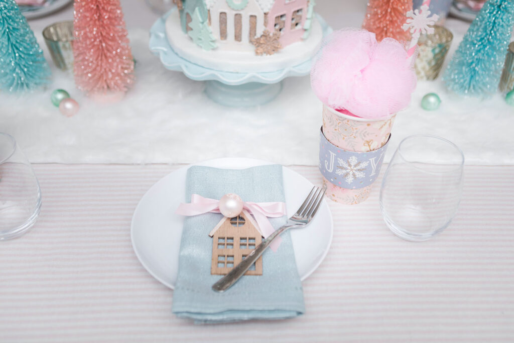 Pink Christmas party place setting. There's a white plate with a silvery blue napkin placed on top. The napkin is wrapped with a pink satin bow and has a small ornament and house cut out attached. To the left is a wine glass and a pink holiday cup favor.