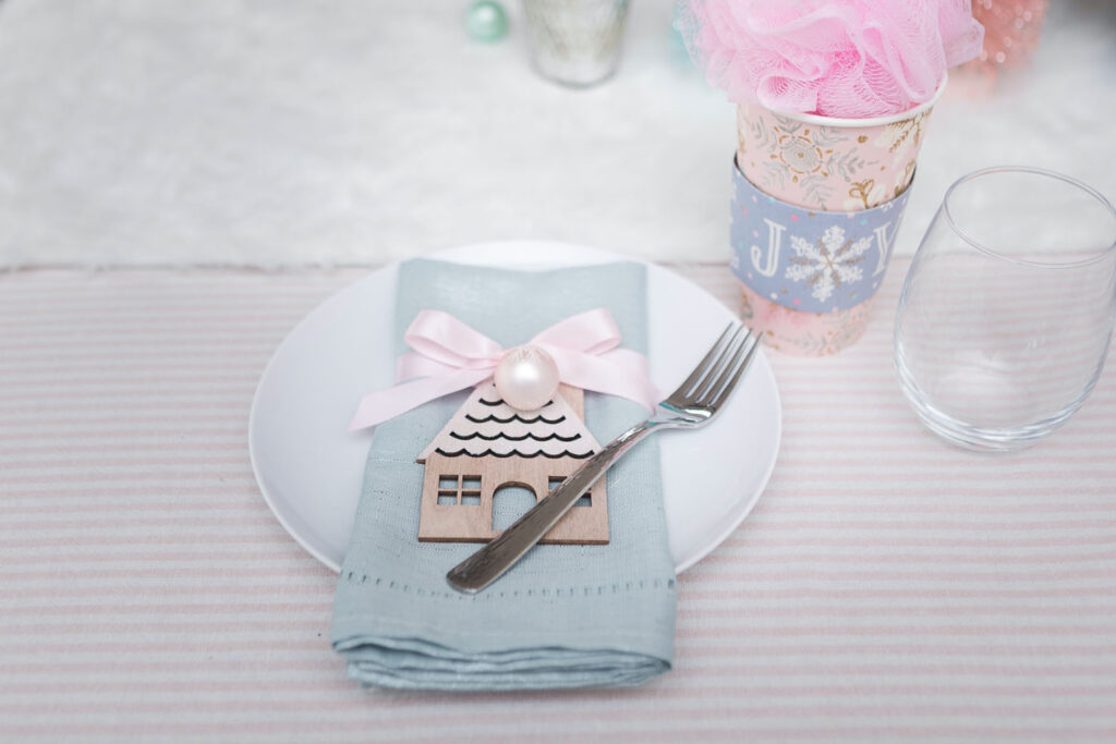 Pink Christmas party place setting. There's a white plate with a silvery blue napkin placed on top. The napkin is wrapped with a pink satin bow and has a small ornament and house cut out attached. To the left is a wine glass and a pink holiday cup favor.