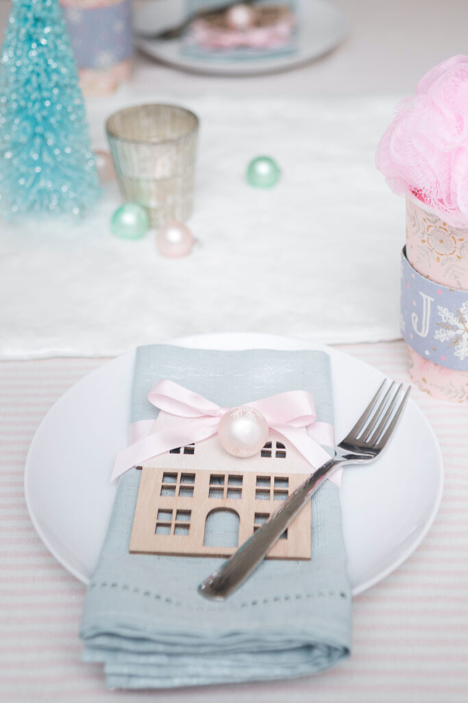 Close up of place setting. There's a white plate with a silvery blue napkin folded on top. The napkin is wrapped with a pink satin ribbon. Attached is a pink mini ornament and and a house cut out.