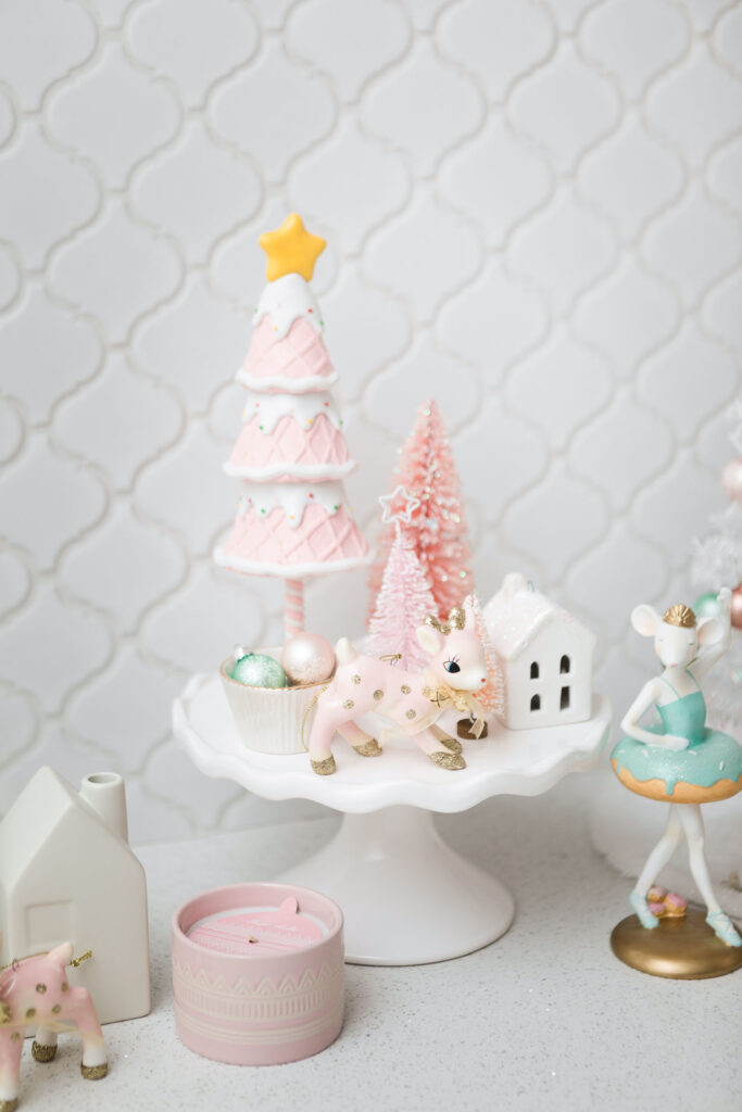 Close up of pink Christmas holiday decor. There's a cake stand topped with a ice cream cone tree, pink deer ornament, ceramic house and pink bottle brush trees.