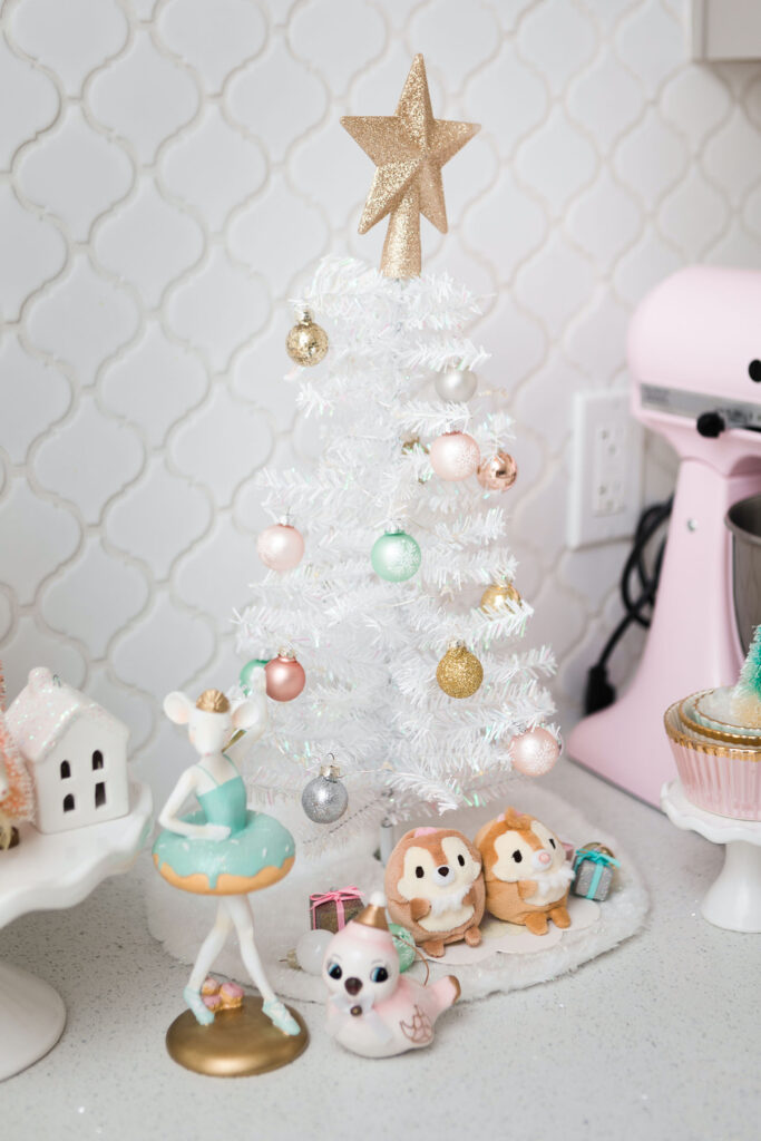 White table top Christmas tree with gold star on top. It's decorated with pink, mint green, gold and silver ornaments. At the base of the tree are miniature chipmunk plushes, a ceramic bird and a mouse ballerina figurine.