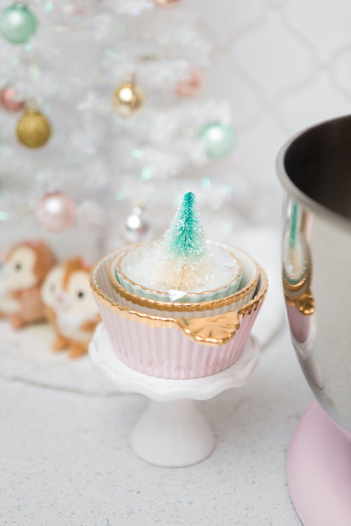 A stack of pink, white and and light green decorative baking cups propped on a tiny cake stand. Inside is faux snow and a mint green bottle brush tree.