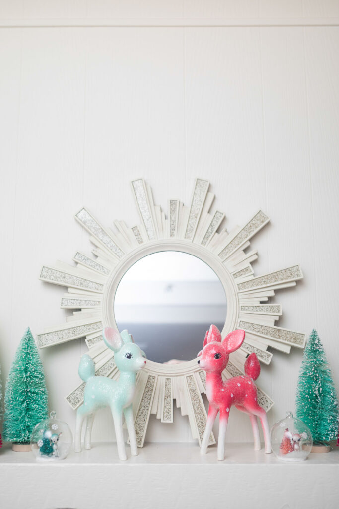 Close up of the top of the fireplace mantle. A cream starburst mirror is placed in the center. A mint and a hot pink deer are placed in the front with mint bottle brush trees to the side.