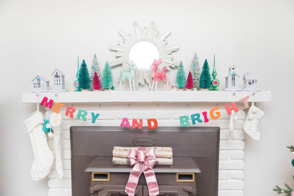 Close up of the top of fireplace mantel. It's a off white brick fireplace topped with a starburst mirror, bottle brush trees and a merry and bright banner. There are also house stocking holders holding cream cable knit stockings.