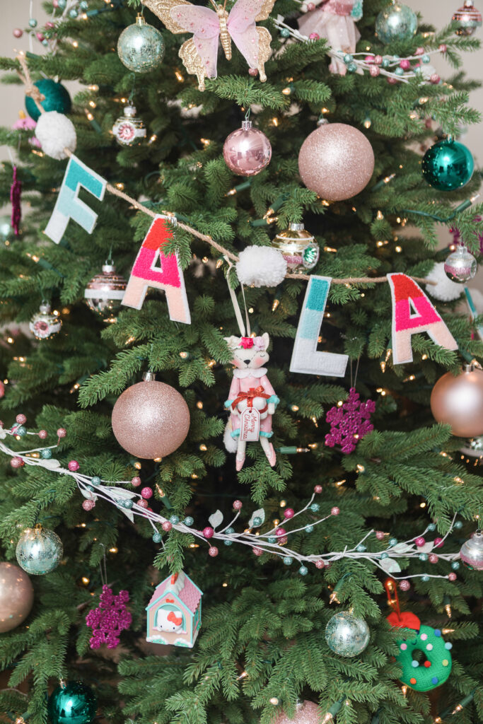 Close up of Christmas tree. There are pink, mint and teal ornaments with a garland that says "Fa la la la" in pink and turquoise letters.