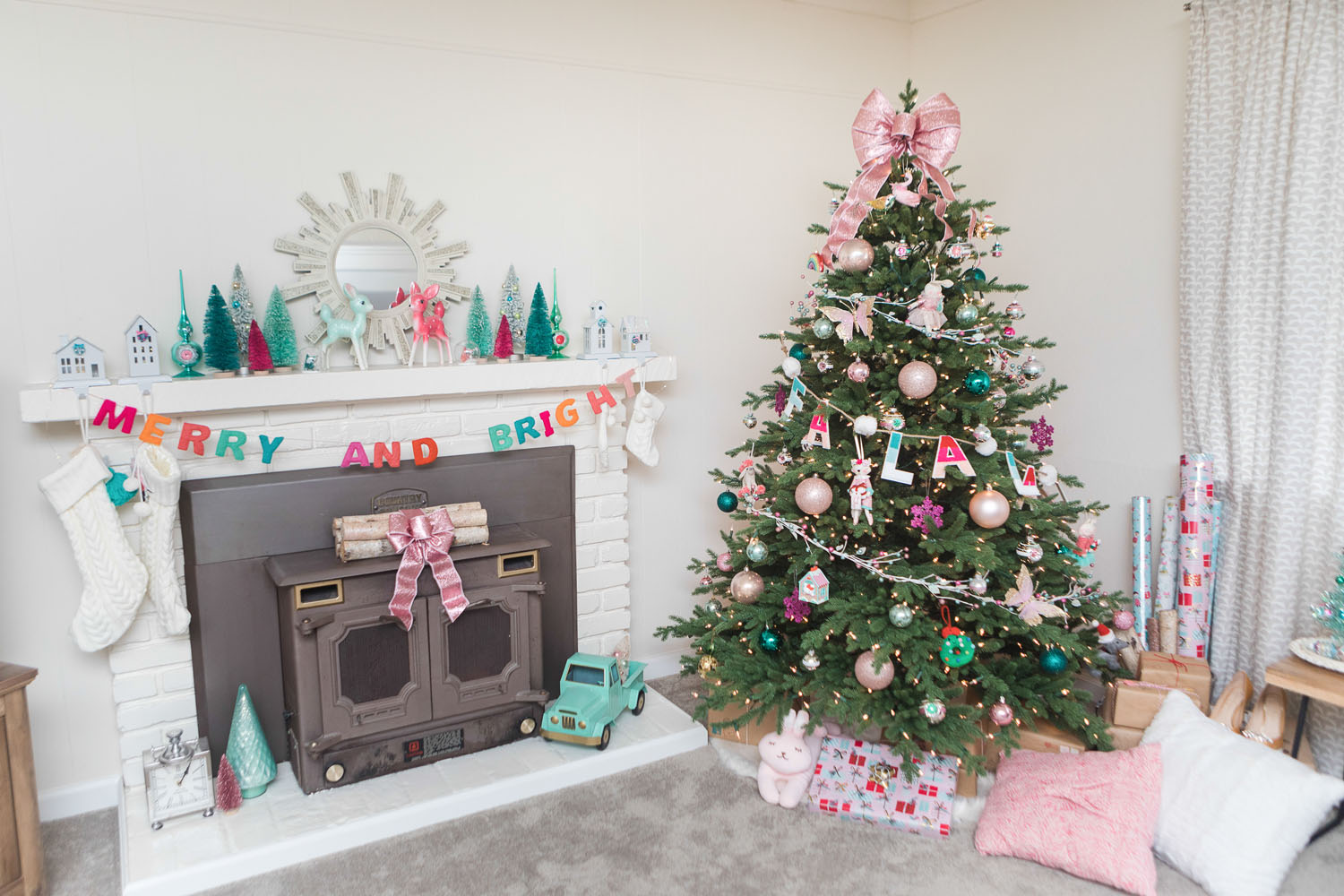Living room with cream walls decorated for Christmas. The fireplace mantel is decorated with colorful bottle brush trees and a "Merry and Bright" garland. The tree is decorated with colorful ornaments and is topped with a giant pink bow.