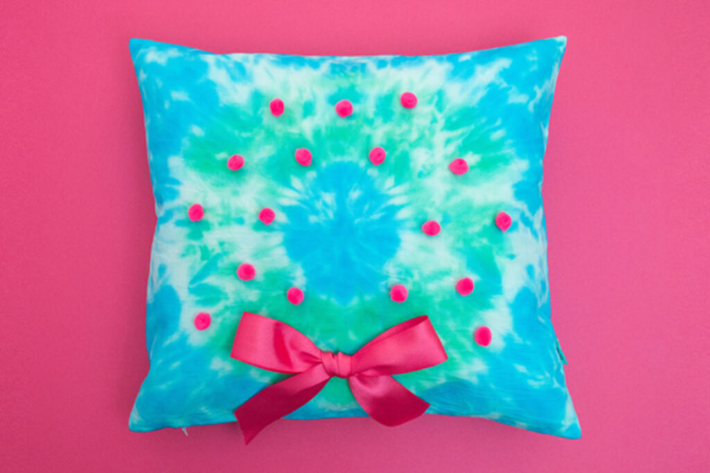 Square pillow cover tie dyed to look like a wreath. It's decorated with pink pom poms and a pink bow.