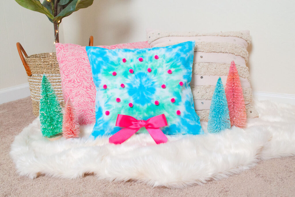 Tie dye wreath pillow amongst other pillows on a faux fur rug. There's a fiddle leaf plant in the back and bottle brush trees placed to the side.