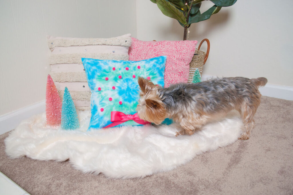 Tie dyed pillow placed amongst two other pillows on a faux fur rug. A yorkshire terrier, Penny, is sniffing the pink bow. 