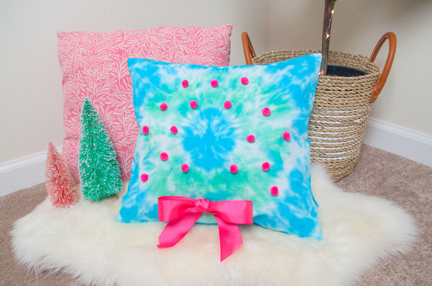 Tie dye wreath pillow placed on faux fur rug. There is a pink pillow and fiddle leaf plant behind it. Bottle brush trees are placed to the left of the pillow.