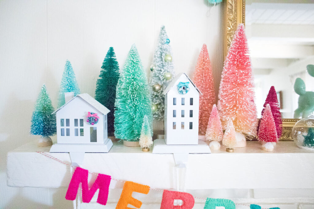 Close up of colorful Christmas mantel. A collection of bottle brush trees arranged in a gradient fashion from teal to mint, to pink, to fuschia. Amongst the trees are two stocking holders in the shapes of houses. There are small wreaths glued to the front.