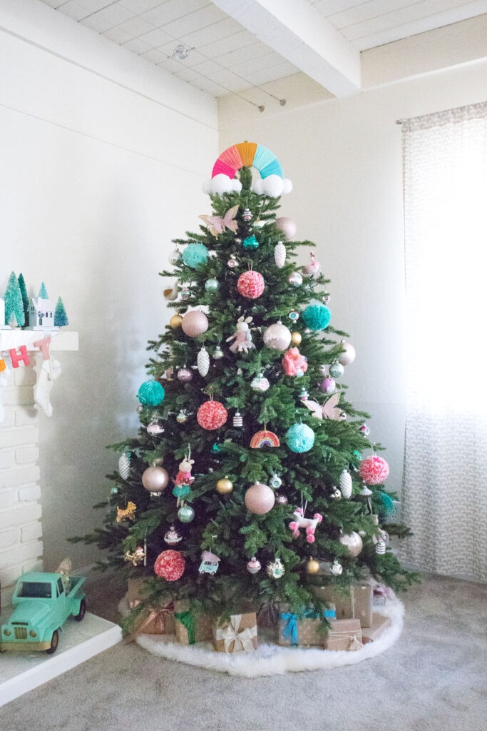 View of Christmas Tree. The topper is a rainbow Blaire made with foam board and yarn. The tree is decorated with ball ornaments, shiny brites, DIY pom poms, and novelty ornaments. The tree skirt is faux fur and gifts wrapped with brown paper are placed under the tree.