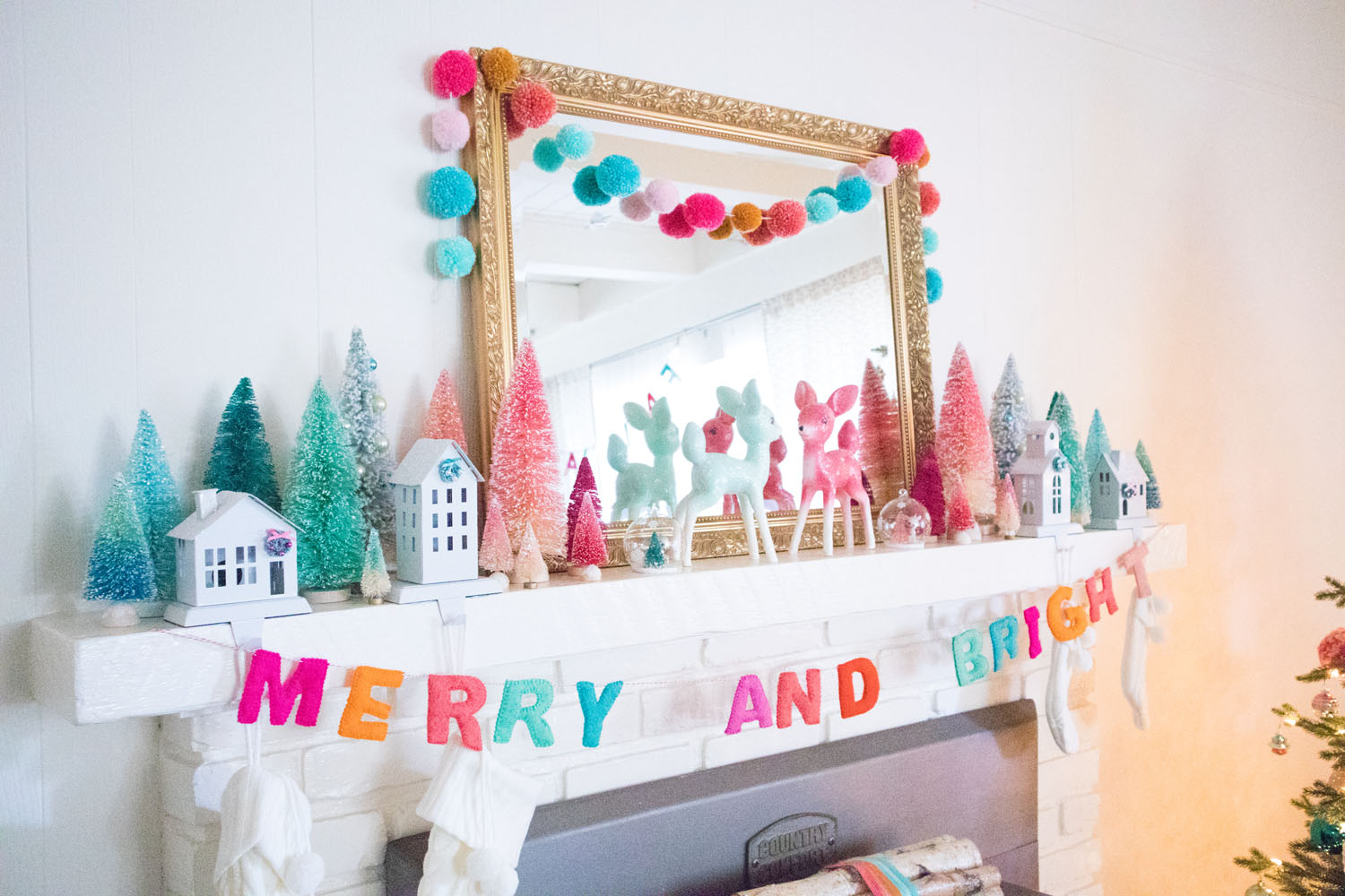 Side view of the fireplace mantel. The mantel is decorated with a large gold mirror, bottle brush trees, deer figurines and colorful garlands. Cream stockings are hung from stocking holders in the shape of houses.