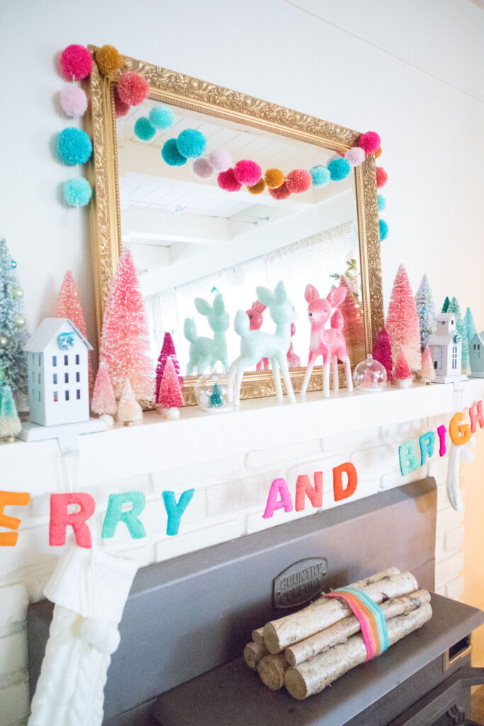 Close up of the fireplace mantel. The focal point is a large gold mirror. In front are two deer figurines and matching globes with mini deer. Next to the deer are bottle brush trees in shades of pink and mint. A pom pom garland is draped over the mirror and a "Merry and Bright" felt garland drapes across the whole mantel.