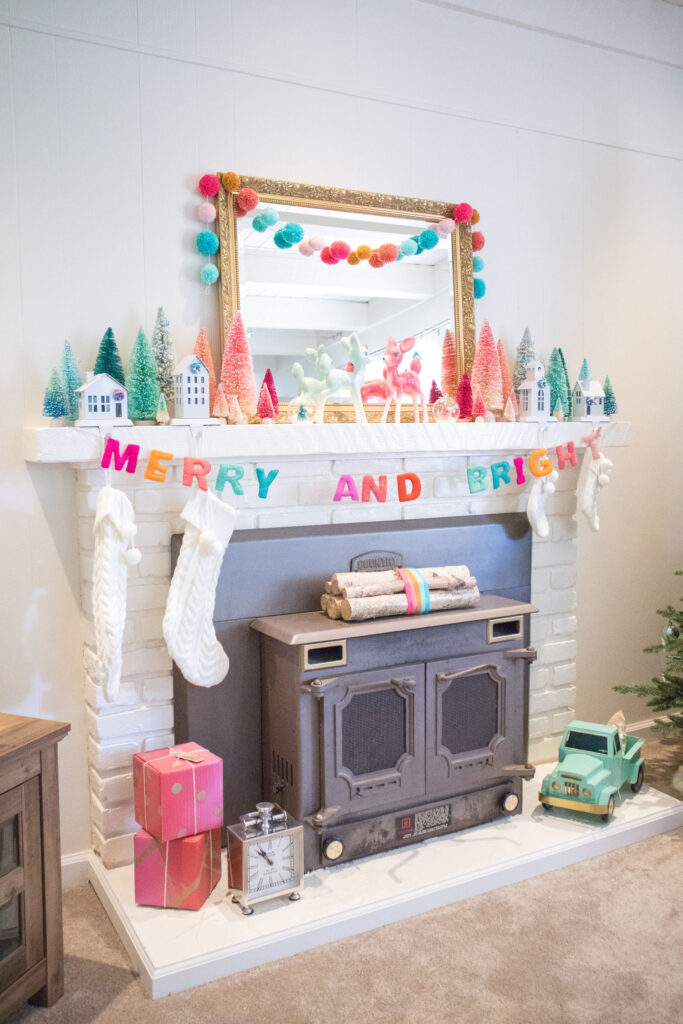 Side view of Blaire's fireplace. The mantel is decorated with a large gold mirror, bottle brush trees, deer figurines and colorful garlands. Cream stockings are hung from stocking holders in the shape of houses. Near the bottom is a bundle of birch logs (tied with rainbow yarn), two red and fuchsia presents and a mint toy truck.