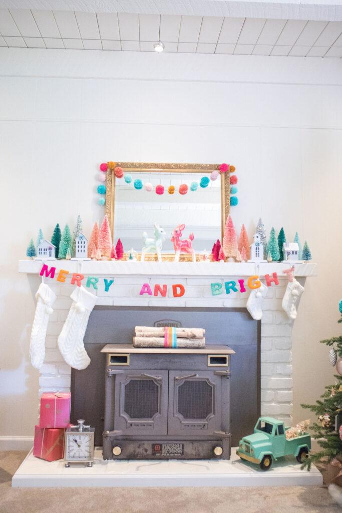 Full view of Blaire's fireplace decor. The mantel is decorated with a large gold mirror, bottle brush trees, deer figurines and colorful garlands. Cream stockings are hung from stocking holders in the shape of houses. Near the bottom is a bundle of birch logs (tied with rainbow yarn), two red and fuchsia presents and a mint toy truck.