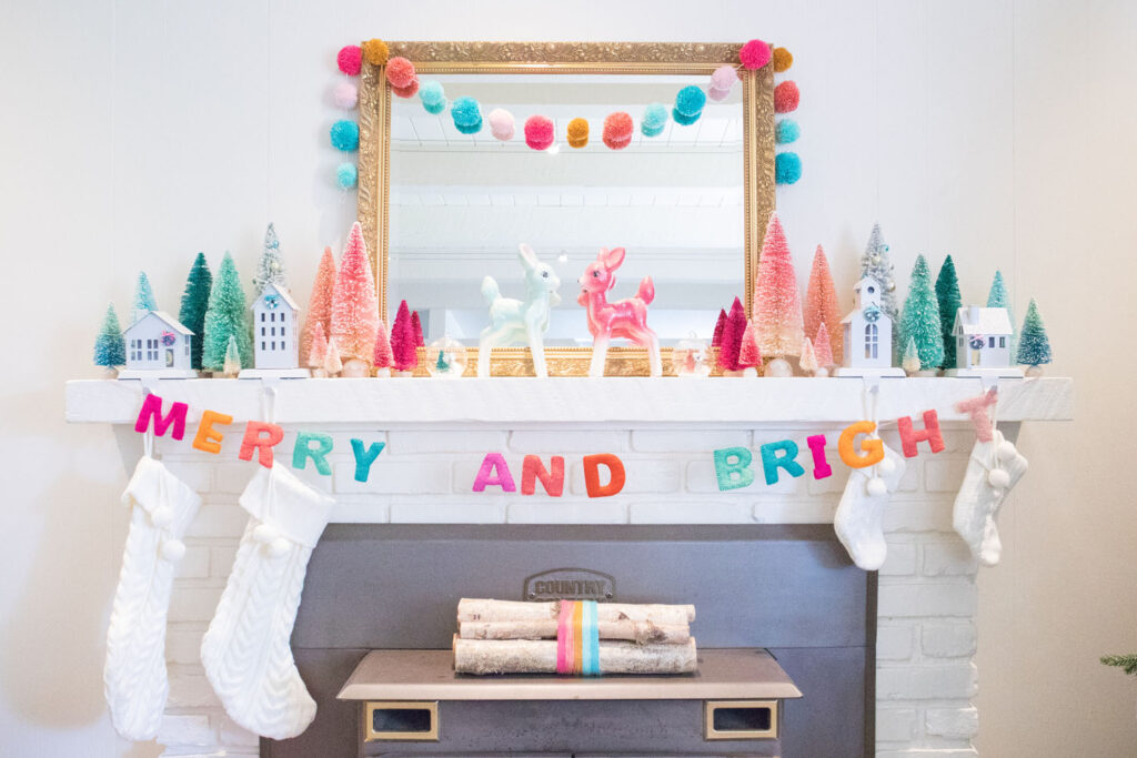 Front view of the fireplace mantel. The mantel is decorated with a large gold mirror, bottle brush trees, deer figurines and colorful garlands. Cream stockings are hung from stocking holders in the shape of houses.