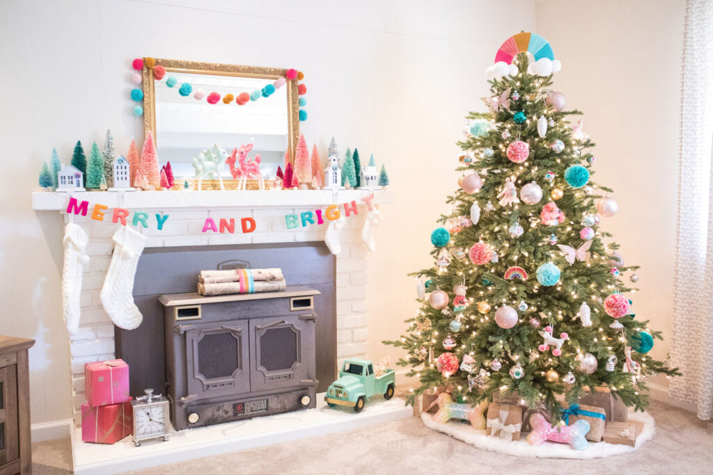 Colorful Christmas living room decor. Fireplace to the left is decorated with a large gold mirror, bottle brush trees, cream stockings and bright garlands. The Christmas tree is filled with whimsical ornaments and pom pom ornaments. The tree is light with warm white lights.The top of the tree has a rainbow tree topper made by Blaire.