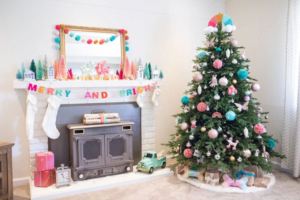 Colorful Christmas living room decor. Fireplace to the left is decorated with a large gold mirror, bottle brush trees, cream stockings and bright garlands. The Christmas tree is filled with whimsical ornaments and pom pom ornaments. The top of the tree has a rainbow tree topper made by Blaire.