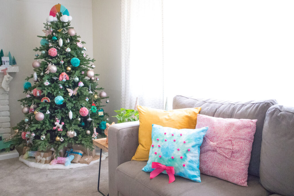 Photo of living room with colorful Christmas tree in the background. On the taupe couch are three colorful pillows - a gold one, a pink printed one with a bow, and a tie dye wreath pillow (made by Blaire from Freshly Fuji).