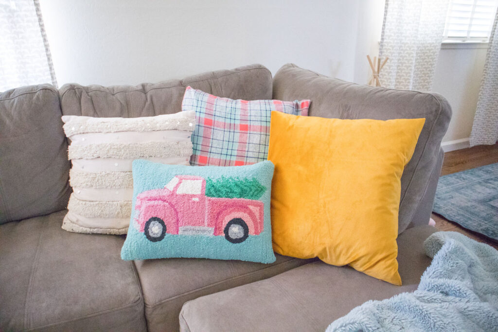 Corner of taupe sectional filled with colorful Christmas pillows - white woven one, gray and coral plaid one, a gold velvet one and a blue one with a pink Christmas truck.